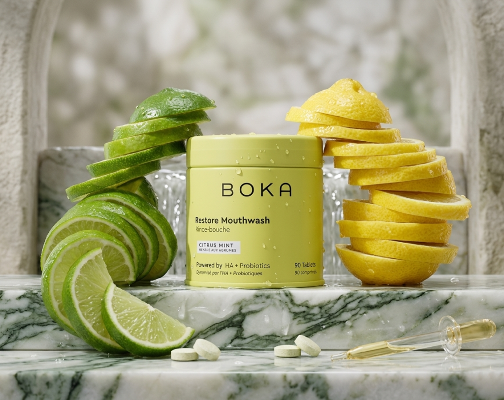 A yellow container of Boka mouthwash tablets in Citrus Mint flavor, flanked by stacked slices of fresh green limes and yellow lemons.