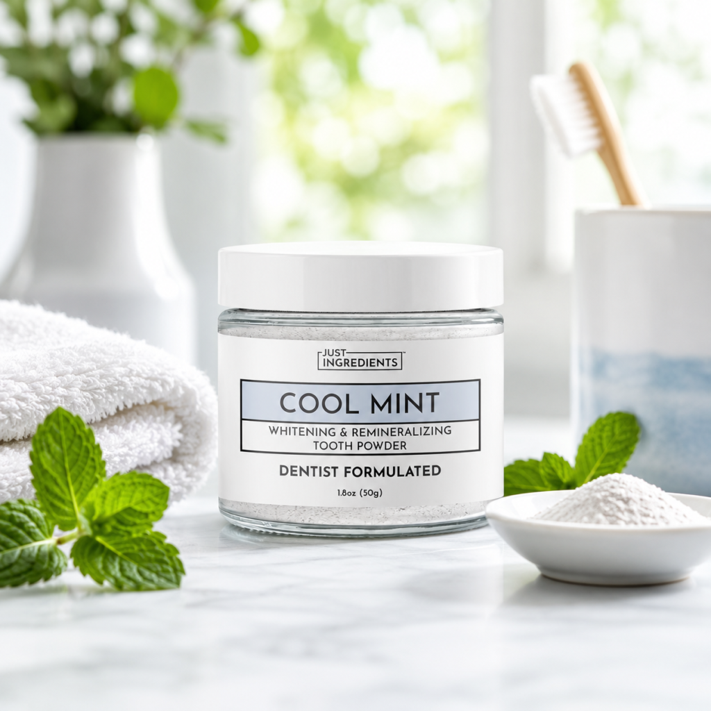 Just Ingredients remineralizing tooth powder displayed with fresh mint leaves, a bamboo toothbrush, white towels, and tooth powder in a calm bathroom setting.