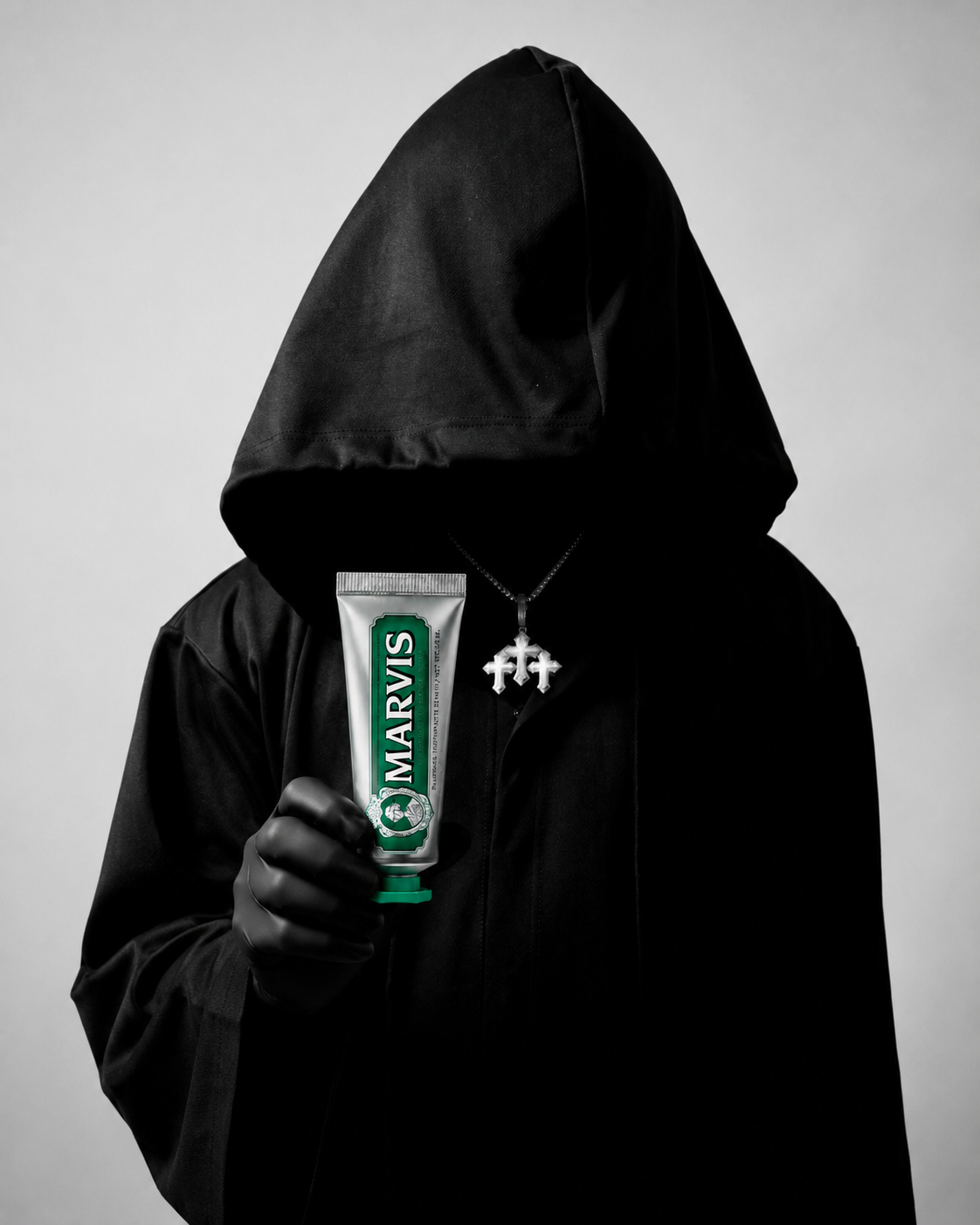 Hooded cult member in a black robe holding a green Marvis toothpaste tube against a light gray background.